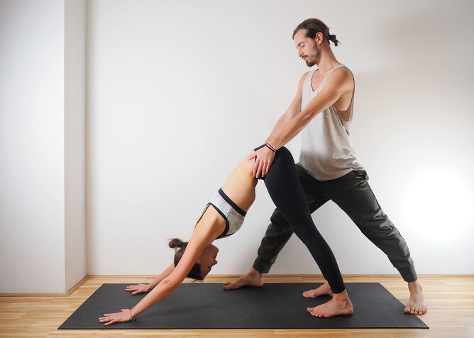 Der herabschauende Hund mit Partner | © SONNENTOR Auf dem Foto sieht man eine junge Frau in der Yogaposition Der herabschauende Hund. Ihr Yogapartner unterstützt sie dabei. | © SONNENTOR